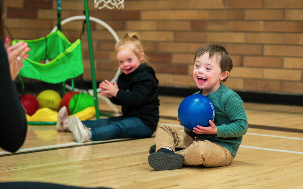 A young boy with intellectual disabilities sits on the gym floor next to another child, smiling while holding a blue rubber ball.