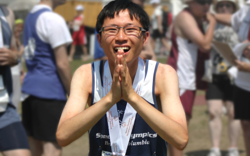 Special Olympics BC athlete with a medal around his neck, looks directly to the camera with hands clasped in front to say thank you.