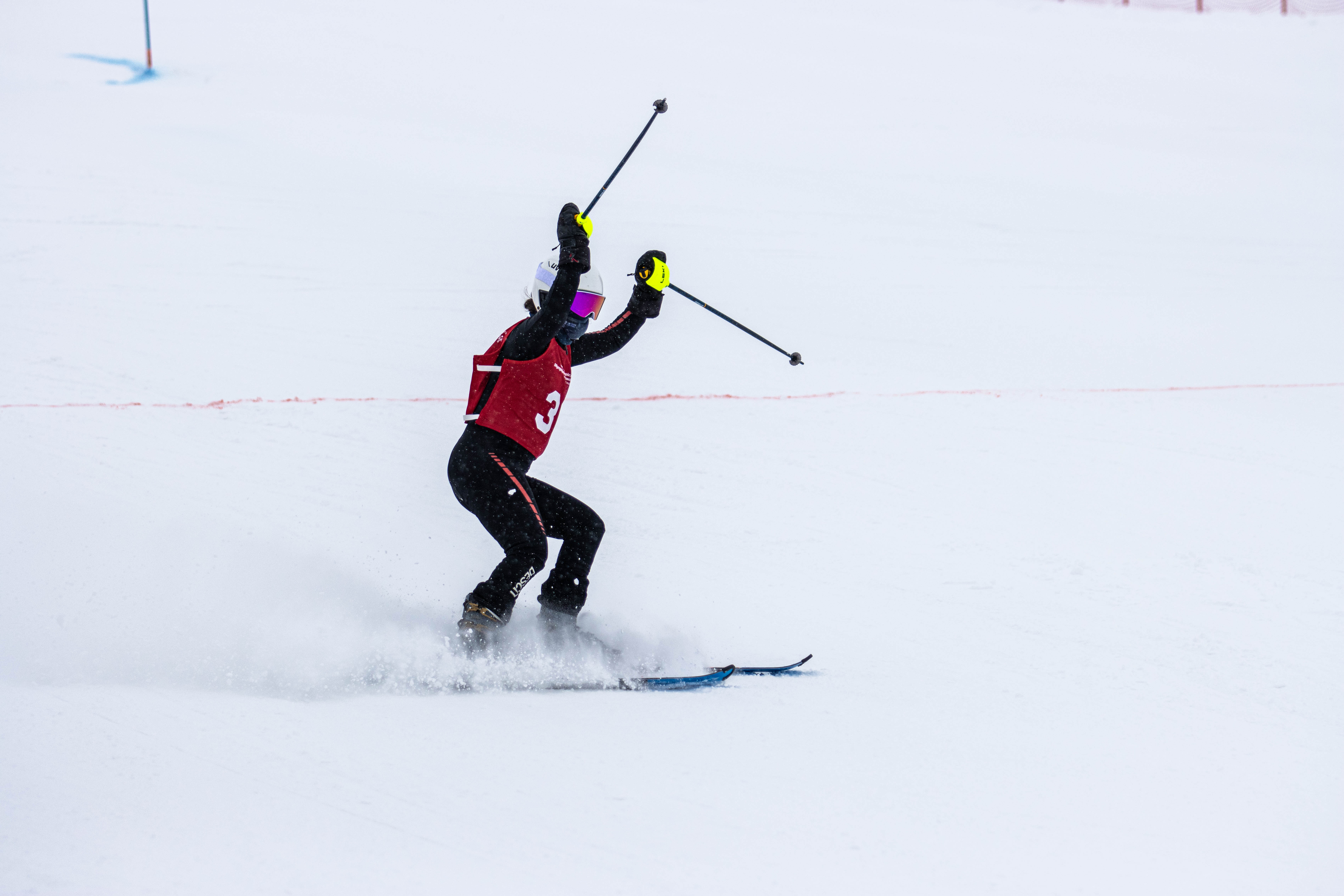 Special Olympics BC alpine skiing athlete celebrates at the finish line with their hands and poles raised in the air and a cloud of snow off their skis as they come to a stop. Special Olympics BC alpine skiing athlete celebrates at the finish line with their hands and poles raised in the air and a cloud of snow off their skis as they come to a stop.