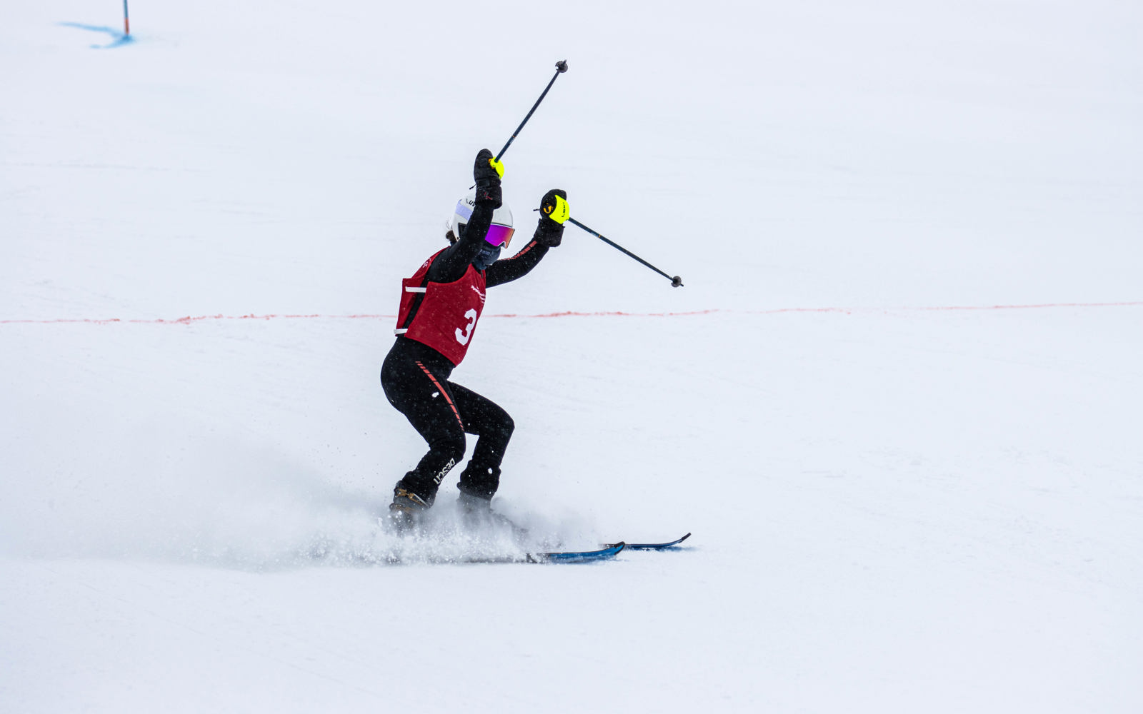 Special Olympics BC alpine skiing athlete celebrates at the finish line with their hands and poles raised in the air and a cloud of snow off their skis as they come to a stop.