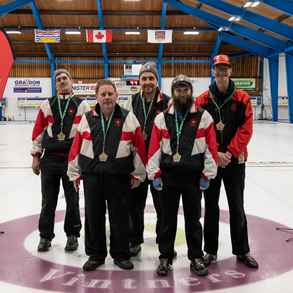 A proud team of Special Olympics BC curling athletes poses together with their coach with medals around their necks.